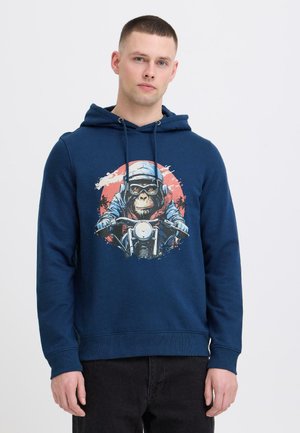 Jonge man draagt een marineblauwe hoodie met een afbeelding van een chimpansee die op een motorfiets rijdt tegen een rode cirkelvormige achtergrond.