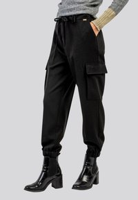 Pantaloni cargo neri con cintura regolabile, tasche laterali e polsini a costine. Indossati con stivaletti neri lucidi con tacco.