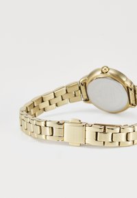 Gold metal link bracelet with a clasp featuring an embossed logo, attached to the back of a wristwatch.