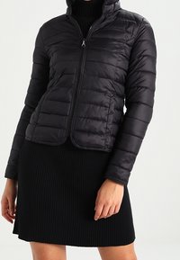 Black padded jacket with a high collar, front zip closure, and quilted texture. Worn over a black ribbed dress.