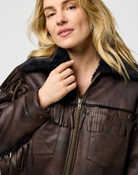 Veste en cuir marron avec des accents en franges, col en fausse fourrure noire, fermeture éclair à l'avant et deux poches à rabat. Surface texturée avec un grain visible.