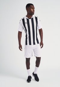 Zwart-wit gestreepte voetbalshirt met korte mouwen, gecombineerd met witte shorts. Zwarte sneakers en witte sokken maken de outfit compleet.