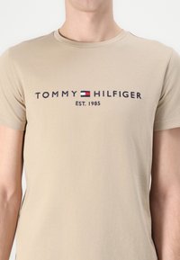 Bežinis medvilnės marškinėliai su "TOMMY HILFIGER" logotipu tamsiai mėlyna spalva, apvadu raudona ir baltos spalvos stačiakampiu, apvaliu kaklu ir trumpomis rankovėmis.
