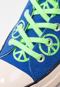 Blue canvas sneaker featuring neon green laces and peace sign graphics. Design includes a glossy cream toe and black rubber sole.