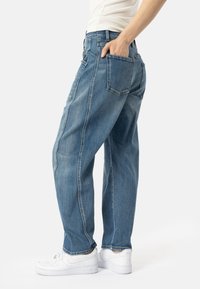 Šviesiai mėlynos džinsinės kelnės laisvai pritaikytos, su šoninėmis siūlėmis, penkiomis kišenėmis ir tiesaus kirpimo dizainu, derinamos su baltomis sportinėmis bateliais.