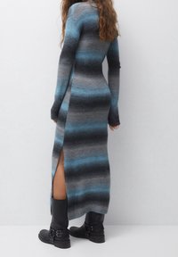 Robe maxi en tricot avec des rayures bleues et grises, manches longues, fente sur le côté et silhouette ajustée. Modèle portant des bottes noires jusqu'aux genoux.