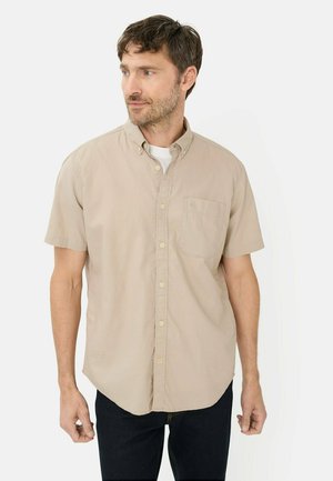 Mand iført beige kortærmet skjorte over hvid t-shirt, stående mod en ensfarvet hvid baggrund, kiggende mod venstre.
