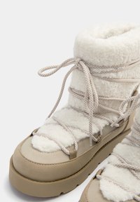 Botas beige con cordones que presentan un forro suave y esponjoso en blanco, material similar al ante, suela gruesa y cordones de patrón intrincado.