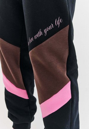 Schwarze Hose mit braunen und rosa geometrischen Patches sowie kursivem rosa Text auf dem Oberschenkel, der "Lebe dein Leben" liest.