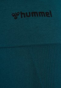 Tkanina w kolorze teal o ribbowej teksturze, z czarnym logo "hummel". Zbliżenie podkreśla gładki, elastyczny materiał oraz bezszwową konstrukcję.