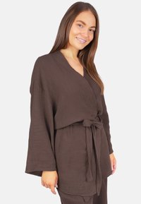 Lächelnde Frau, die ein locker sitzendes, braunes, strukturiertes Wickeloberteil mit weitem Ärmel trägt, das an der Taille gebunden ist, vor einem schlichten hellen Hintergrund stehend.