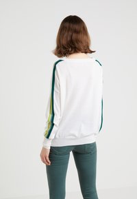 Maglione bianco a maniche lunghe con strisce verdi e gialle sulle spalle. Texture morbida e vestibilità rilassata, abbinato a pantaloni turchesi.