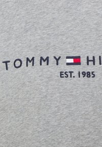 Tissu gris avec texte brodé bleu marine "TOMMY HILFIGER" présentant un logo rectangulaire rouge, blanc et bleu marine et "EST. 1985" en dessous.