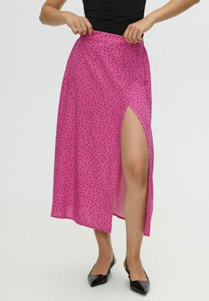 Mujer que lleva una falda midi rosa con pequeñas manchas negras, abertura lateral alta, top negro sin mangas y zapatos negros con punta.