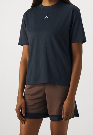 Persoon draagt een donker T-shirt met korte mouwen met een klein wit Jumpman-logo en bruine sportshorts met donkere bies, staand tegen een effen achtergrond.