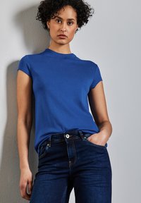 Modrá triko s krátkým rukávem a kulatým výstřihem, kombinované s tmavými džínami klasického střihu a s knoflíkovým zapínáním.