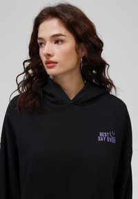 Sudadera negra con capucha amplia, corte holgado y gráfico morado "¡MEJOR DÍA DE LA HISTORIA!" en el frente. Fabricada con un tejido suave y texturizado.