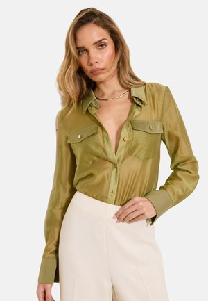 Frau trägt olivgrüne Satinbluse mit Knöpfen und Brusttaschen und cremefarbene Hose mit hoher Taille, posiert mit einer Hand an der Hüfte.