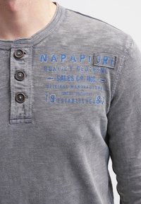 Nahaufnahme einer Person, die ein graues Langarmshirt mit drei braunen Knöpfen und blauem Drucktext auf der Brust und der Schulter trägt.