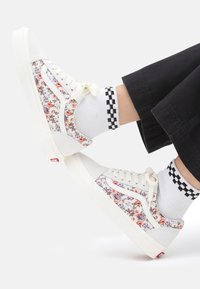 Sneakers bianchi con stampa floreale, caratterizzati da un design low-top, chiusura con lacci e suole testurizzate. Abbinati a calzini a quadretti.