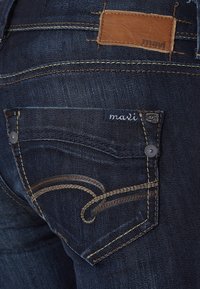 Los jeans de mezclilla azul oscuro presentan un bolsillo trasero con una marca de cuero en color marrón, costuras decorativas en un patrón de espiral y acentos de botones metálicos.