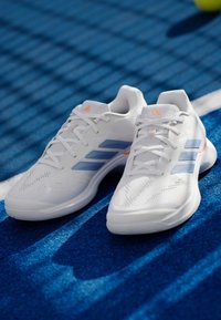 Άσπρα αθλητικά παπούτσια με υφή στην επάνω επιφάνεια, διάτρητα, με γαλάζιες λεπτομέρειες και πορτοκαλί διακοσμητικά στη φτέρνα. Κατάλληλα για αθλητικές δραστηριότητες.