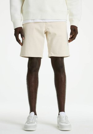 Man wearing cream-colored shorts, white sneakers, and a white sweatshirt, standing with hands relaxed by his sides.