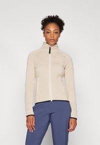 Beige bunda na zip s vysokým límcem, kontrastní černé detaily na manžetách a lemu, vyrobená z texturované tkaniny. Model má na sobě námořnické kalhoty.