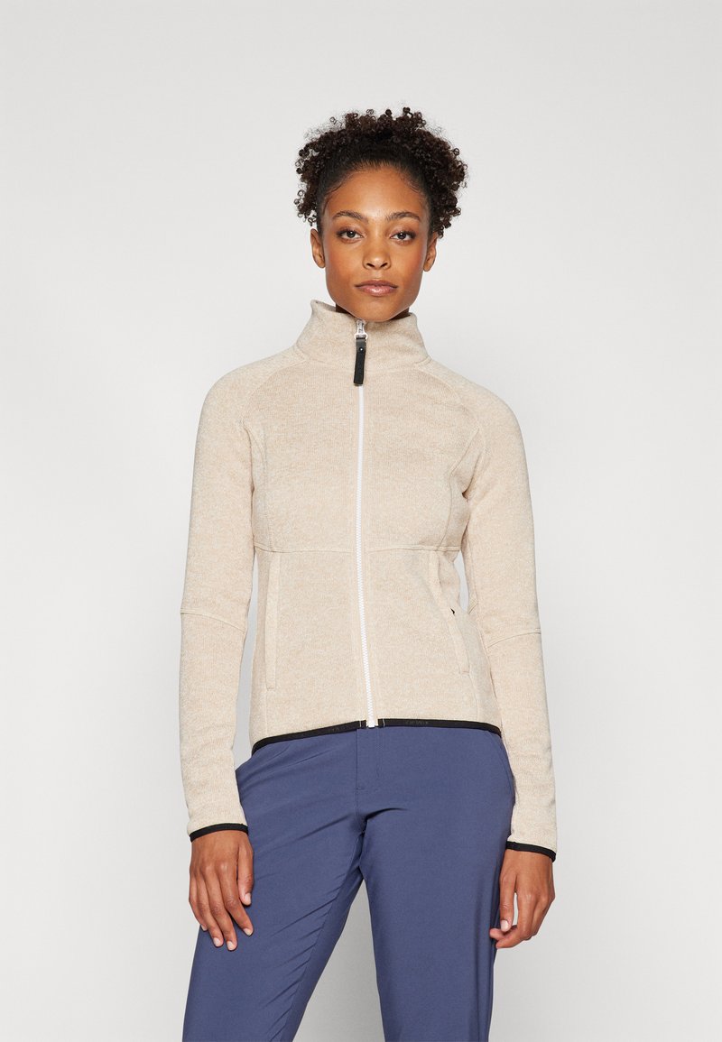 Beige bunda na zip s vysokým límcem, kontrastní černé detaily na manžetách a lemu, vyrobená z texturované tkaniny. Model má na sobě námořnické kalhoty.