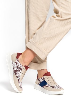 Person wearing beige corduroy pants and beige, white, burgundy, and blue patterned sneakers with white soles, standing on white background.
