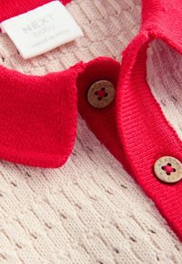 Pull bébé beige en tricot avec col rouge et boutons estampillés « NEXT » ainsi qu’une étiquette indiquant « NEXT baby MADE IN INDIA ».