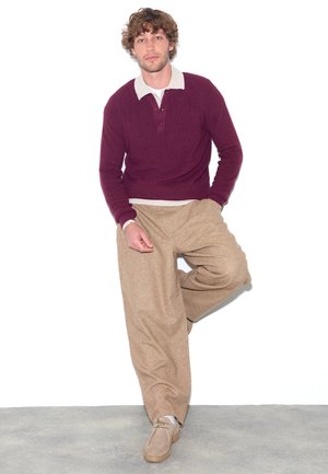 Junger Mann mit lockigem Haar, der einen burgunderfarbenen Pullover über einem weißen Hemd mit Kragen, beige weite Hosen und braune Schuhe trägt, steht vor einer weißen Wand.