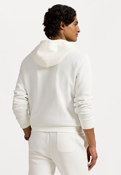 Fehér kapucnis pulóver bordázott mandzsettákkal, sima textúrával és kenguruzsebből. Az összhangban lévő fehér nadrággal viselve, egyszerű hátul nézve.