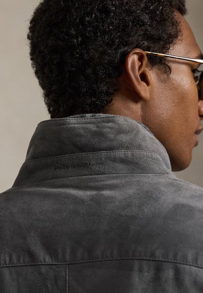 Γκρι σουέντ σακάκι με ψηλό γιακά, με διακριτικό λογότυπο στο πίσω μέρος. Η υφή είναι απαλή και λεία, αναδεικνύοντας την ποιότητα του υλικού.