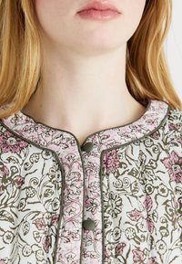 Material floral, alb, cu accente roz și verde; decolteu rotund cu detalii contrastante și nasture legat.