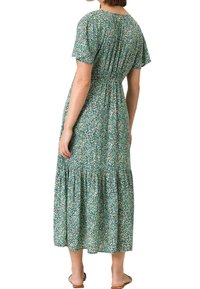 Femme portant une robe midi verte à fleurs avec manches courtes et taille froncée, debout de dos à la caméra.