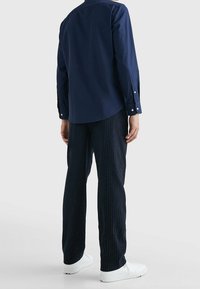 Hombre con una camisa de manga larga azul marino, pantalones oscuros a rayas y zapatillas blancas, de pie frente a un fondo blanco liso.