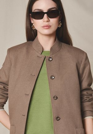 Kobieta w czarnych, prostokątnych okularach przeciwsłonecznych, dużych złotych kolczykach-kołach, brązowej, teksturowanej, zapinanej na guziki kurtce i zielonym topie, stojąca na jednolitym tle.