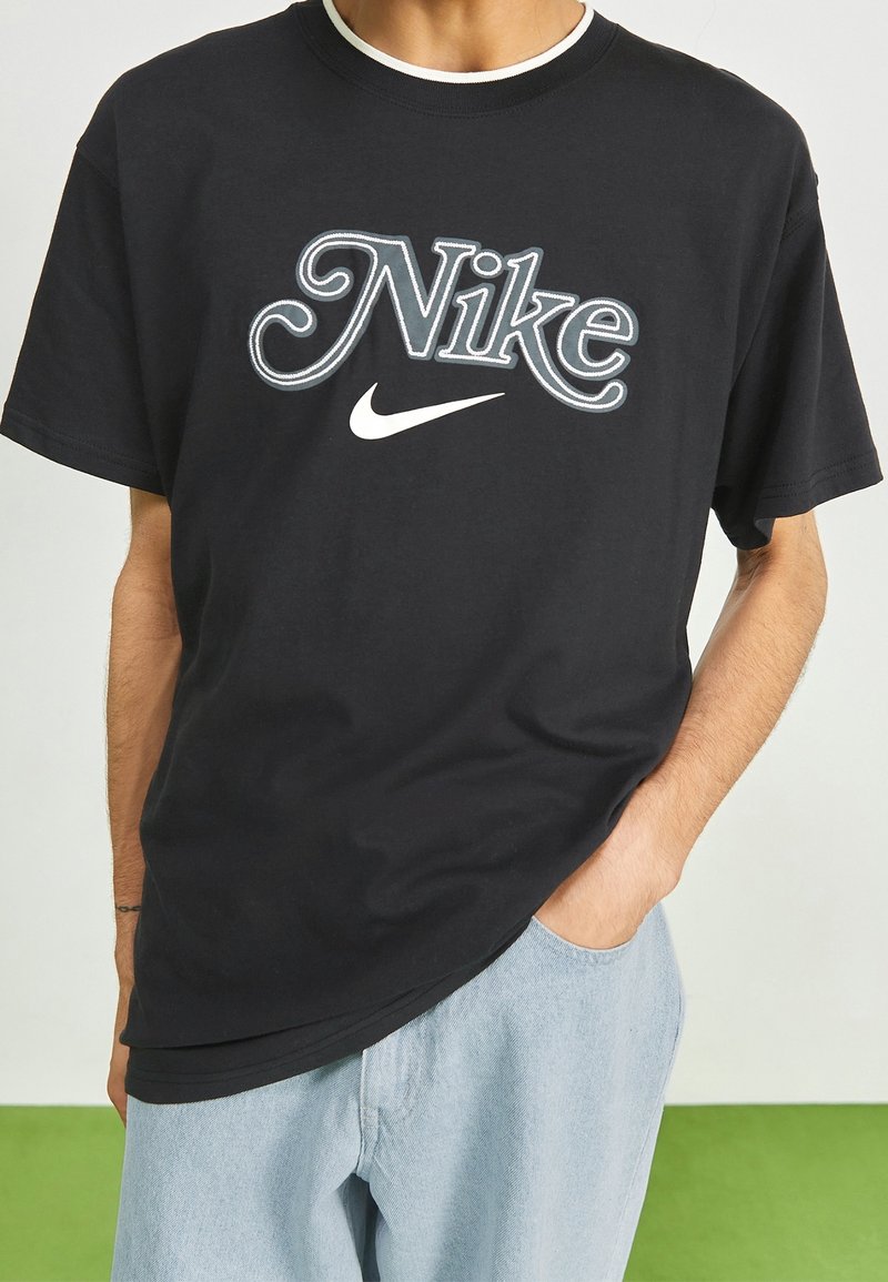 Svart t-shirt i bomull med en stor, stiliserad vit "Nike"-logotyp med swoosh, rund halsringning och lös passform.