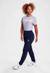 Jasnoszara koszulka z białym logo, zestawiona z granatowymi dresami z bocznym paskiem i elastycznym pasem. Białe buty sportowe.