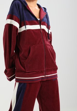 Persoon die een maroon en donkerblauw velours trainingspak met witte strepen en een ritssluiting aan de voorkant draagt, staand tegen een effen lichtgrijze achtergrond.