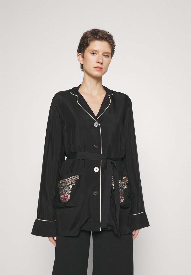 Zwarte satijnen jas met lange mouwen, een ceintuur in de taille, witte piping en decoratieve, kleurrijke patroonzakken.