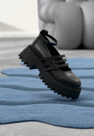 Scarpa platform nera con tre cinturini a fibbia e suola dentellata, posata su un tappeto blu texturizzato con una scultura sfocata sullo sfondo.