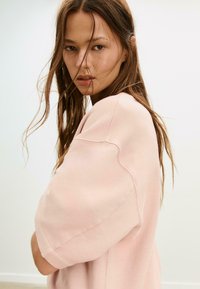 Helles rosa Oversized-Sweatshirt mit weicher Textur. Verfügt über breite, abgerundete Ärmel und einen lässigen Schnitt, vor einem neutralen Hintergrund.