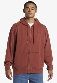 Hnedo-červená zipová mikina vyrobená z jemného materiálu, so prednou vreckom, nastaviteľnými šnúrkami a malým logom na hrudi.