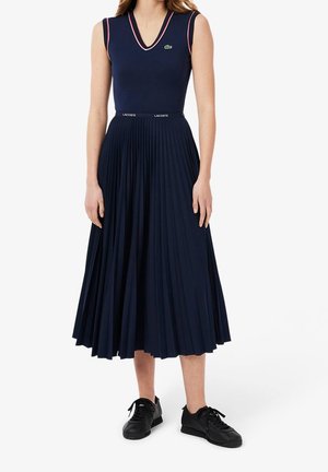 Mujer con top sin mangas azul marino de escote en V con ribete rojo, falda midi plisada azul marino con cinturilla Lacoste y zapatillas negras.