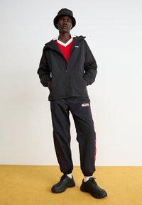 Black zip-up jacket and matching pants, red accents, bucket hat, black chunky sneakers, and a red and white v-neck shirt layered underneath.