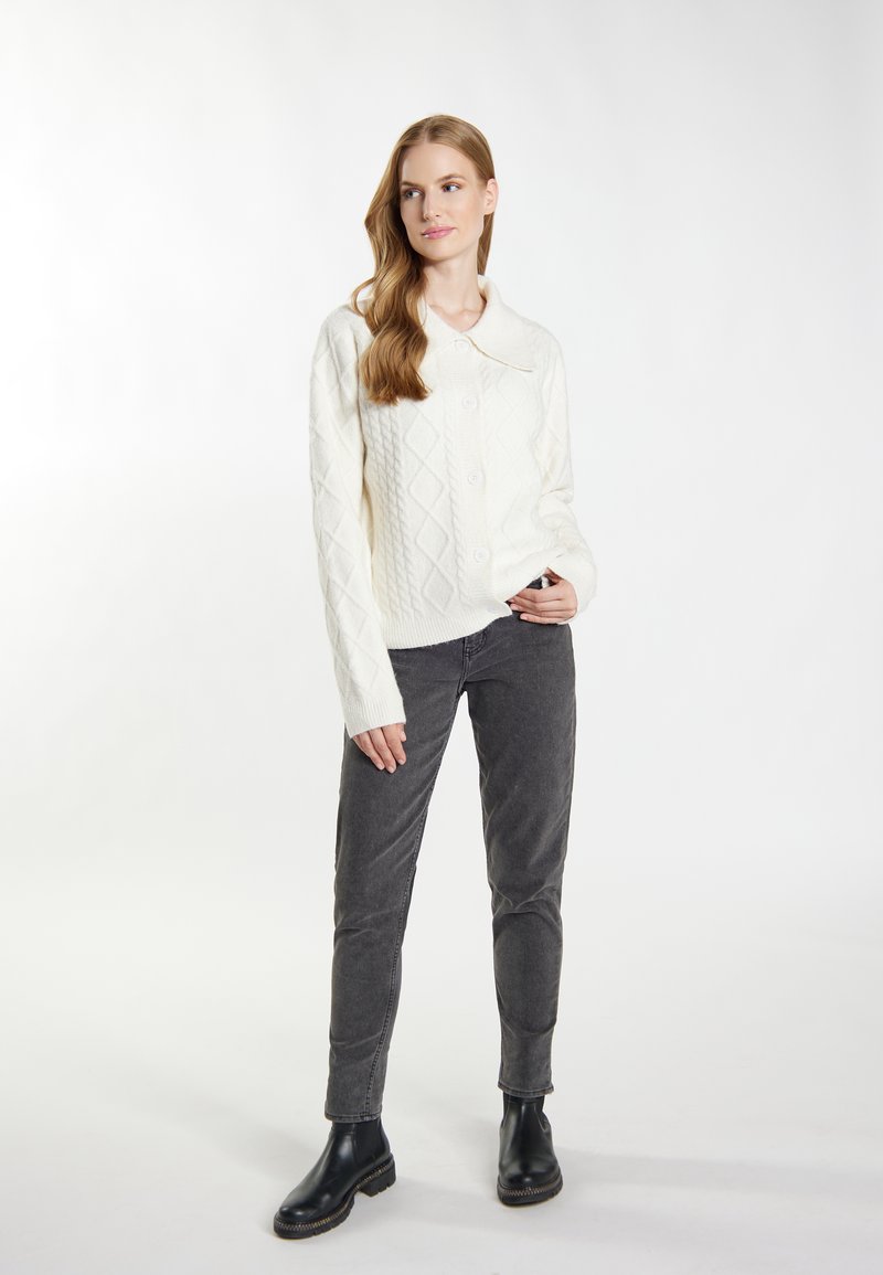 Weißer gestrickter Cardigan mit Rautenmuster, Knopfverschluss und umschlagendem Kragen. Kombiniert mit dunkelgrauen, enganliegenden Hosen und schwarzen Stiefeletten.