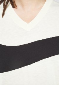 Pull en V en tricot écru avec une large bande noire sur la poitrine, présentant une texture lisse et un col côtelé subtil.