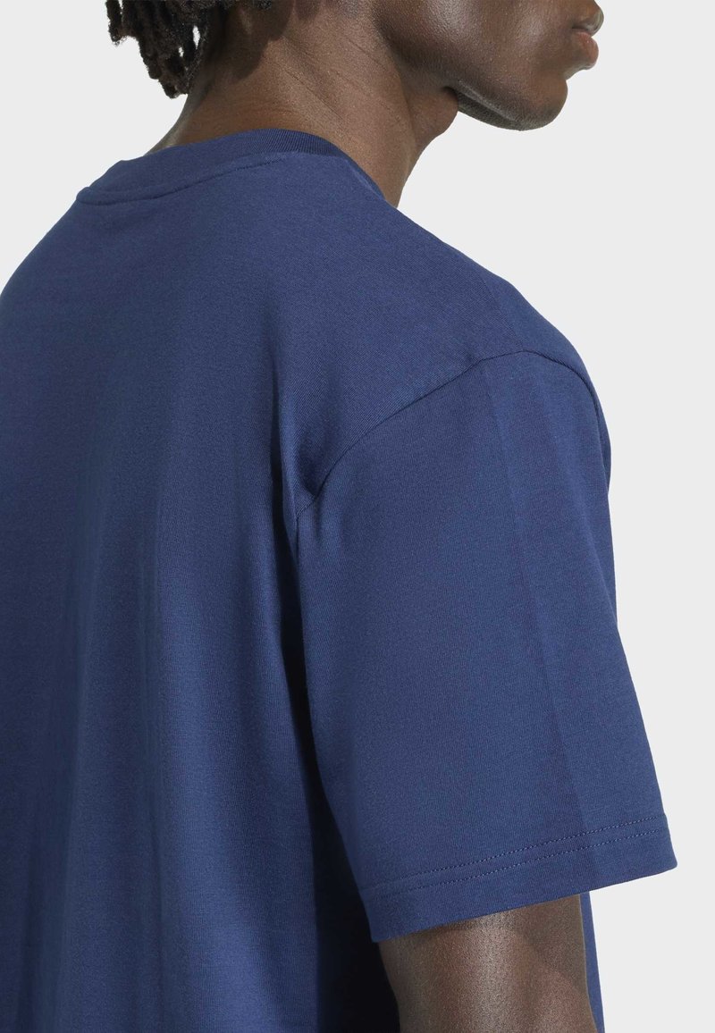 Primo piano di una persona che indossa una maglietta semplice in cotone blu scuro a maniche corte su uno sfondo grigio chiaro.